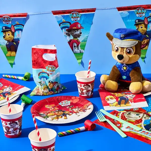 Paw Patrol themed party tableware with character plates, cups, bunting, and a plush toy arranged on a blue background